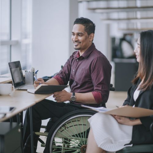Indian white collar male worker in wheelchair having cheerful discussion conversation with his female asain chinese colleague coworking in creative office workstation beside window