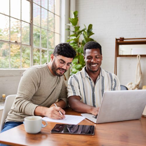 Young multiracial gay couple smiling while going over their home finances together at a table in their living room at home